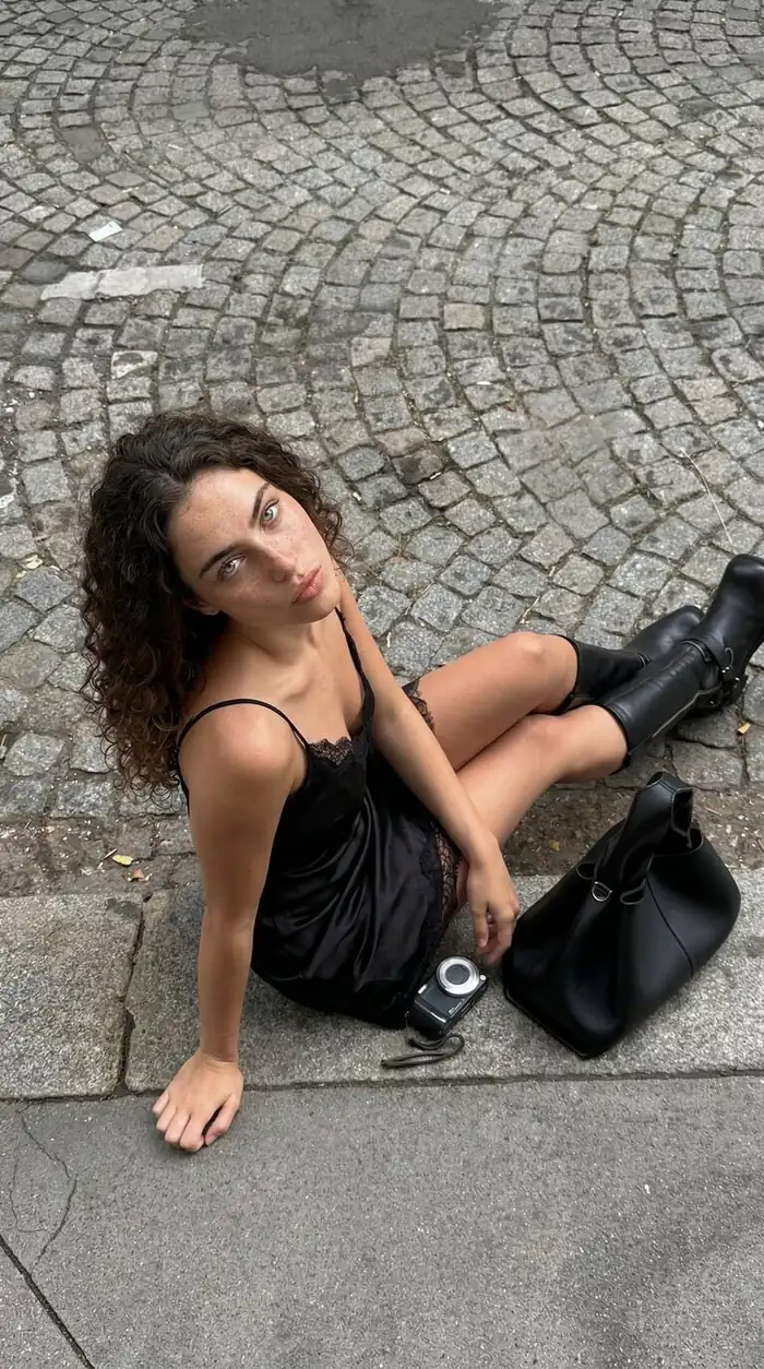Overhead vertical portrait of a woman sitting on a cobblestone street with a black dress, boots, camera, and bag, showcasing urban fashion and outdoor lifestyle.